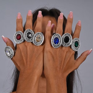 Può includere: Cinque anelli d'argento con pietre preziose colorate. Ogni anello presenta un design decorato con una grande pietra di forma ovale al centro. Le pietre sono rossa, nera, marrone, blu e verde.