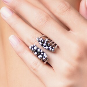 May include: A silver ring with a unique design featuring a cluster of small spheres on each side. The ring is open and adjustable.