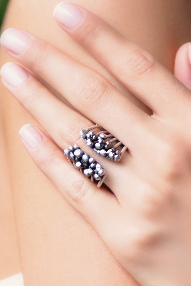 May include: A silver ring with a unique design featuring a cluster of small spheres on each side. The ring is open and adjustable.