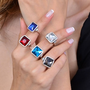 Könnte beinhalten: Fünf Silberringe mit quadratischen Edelsteinen. Die Ringe haben einen blauen, roten, hellblauen, klaren und grauen Edelstein.