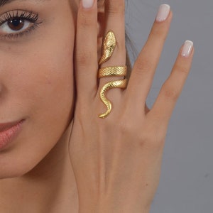 May include: A gold-toned snake ring with a textured finish. The ring is worn on the index finger of a hand.