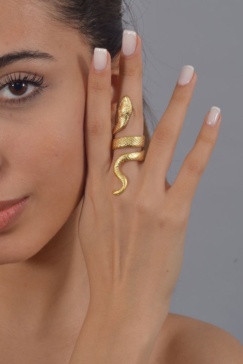 May include: A gold-toned snake ring with a textured finish. The ring is worn on the index finger of a hand.