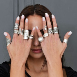 Peut inclure: Des mains de femme portant une variété d'anneaux en argent. Les anneaux sont empilés sur ses doigts et présentent différents motifs, notamment des formes géométriques, des flèches et des surfaces texturées.