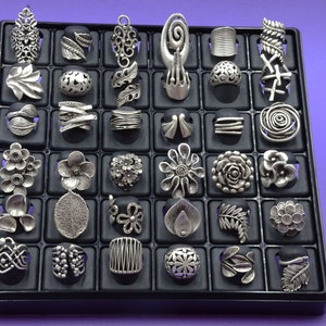 May include: A black display case containing 36 silver rings. The rings are arranged in a grid pattern and feature a variety of designs, including flowers, leaves, and geometric shapes.