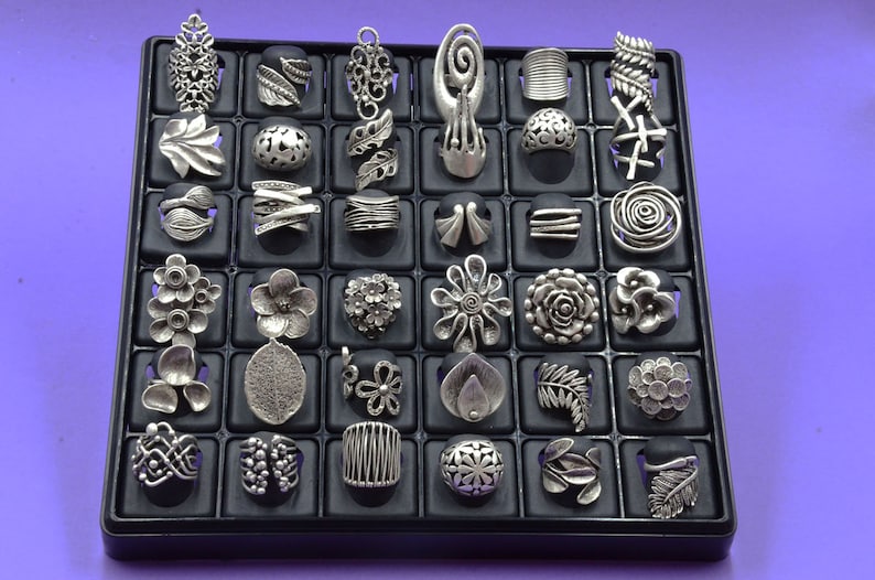 May include: A black display case containing 36 silver rings. The rings are arranged in a grid pattern and feature a variety of designs, including flowers, leaves, and geometric shapes.