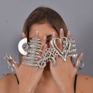 May include: A set of silver-toned rings with various designs, including a heart, a tree, a musical note, and a spiral. The rings are worn on the fingers of a person's hands.