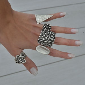 May include: A set of five silver rings with different designs. The rings are stacked on a finger and feature a variety of textures and shapes, including a square, a triangle, a circle, and a spiral.
