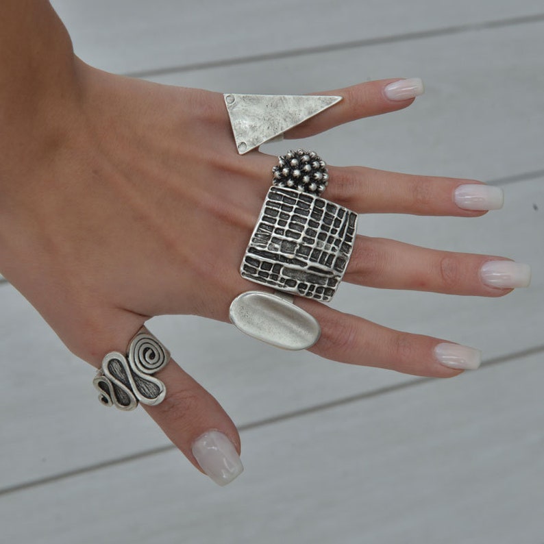 May include: A set of five silver rings with different designs. The rings are stacked on a finger and feature a variety of textures and shapes, including a square, a triangle, a circle, and a spiral.