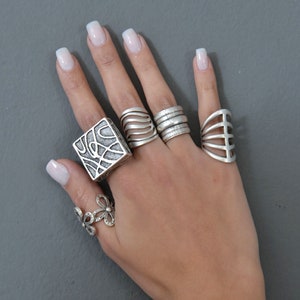 May include: A hand wearing five silver rings. The rings are all different styles, including a square ring with a geometric design, a ring with a wave pattern, a ring with a simple band, a ring with a cage design, and a ring with a flower design.