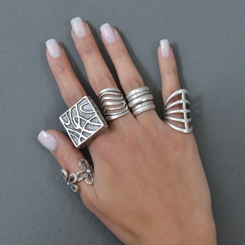 May include: A hand wearing five silver rings. The rings are all different styles, including a square ring with a geometric design, a ring with a wave pattern, a ring with a simple band, a ring with a cage design, and a ring with a flower design.