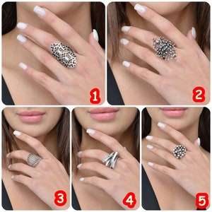May include: Five different silver rings with unique designs. The rings are shown on a model's hand with white nail polish.