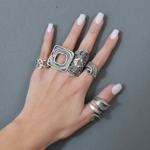 May include: A hand model wearing five silver rings. The rings are all different styles, including a square ring, a floral ring, and a twisted ring.