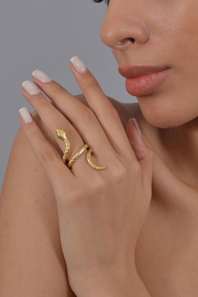 May include: A gold-toned snake ring with a textured finish. The ring is wrapped around a finger and the snake's head is facing upwards.