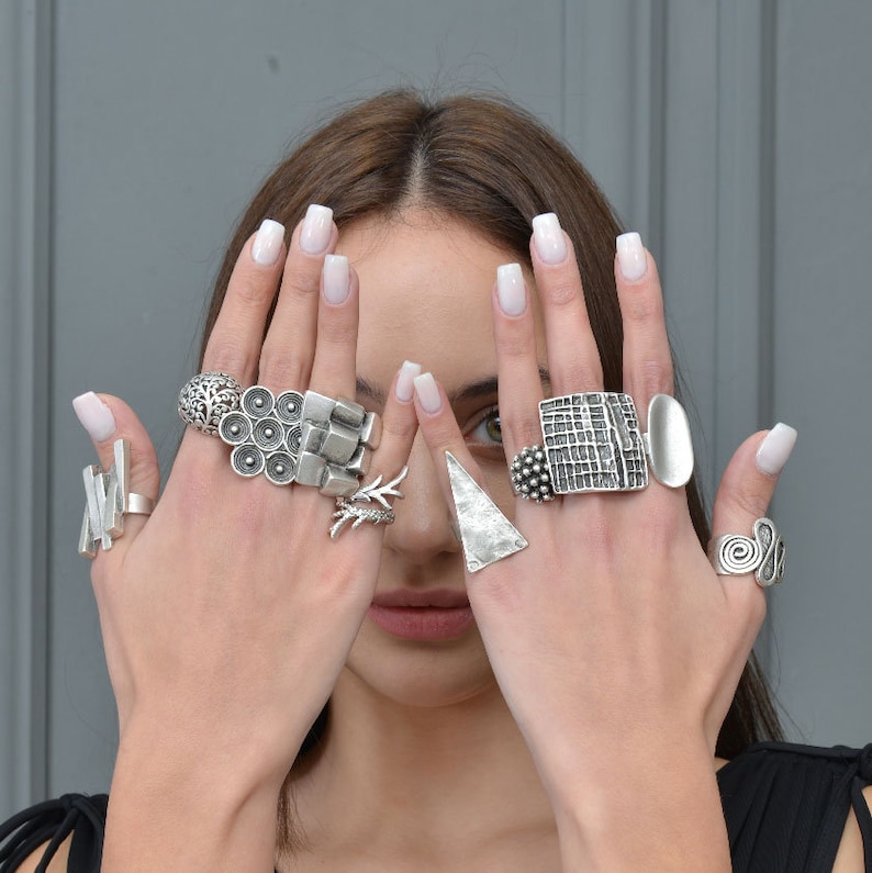 May include: A woman's hands wearing a variety of silver rings. The rings are all different shapes and sizes, and some have intricate designs. The rings are stacked on each finger, creating a bold and stylish look.