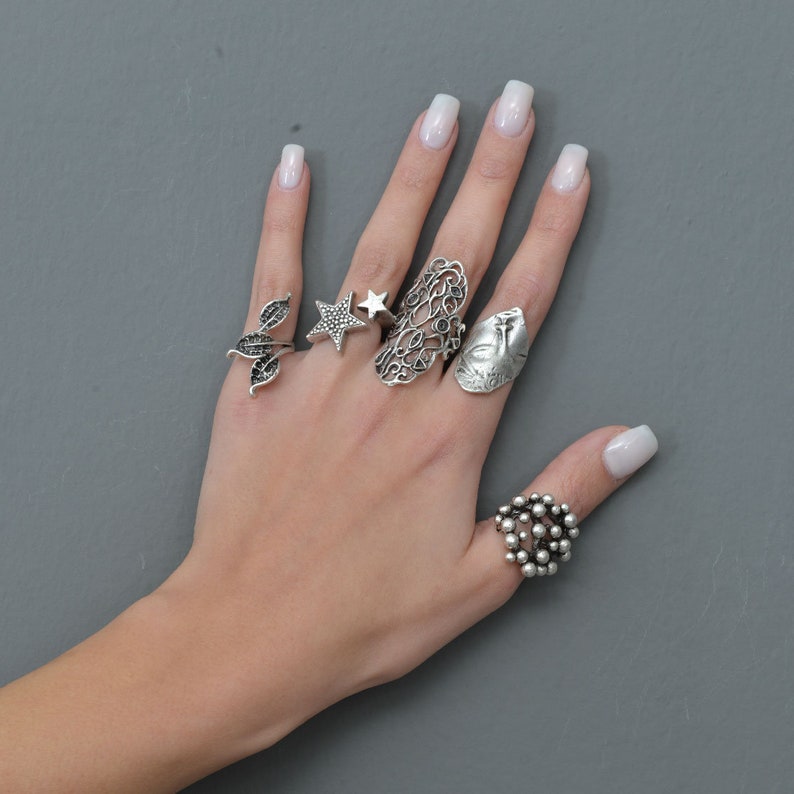 May include: A hand wearing five silver rings. The rings feature a variety of designs, including a star, a leaf, a peacock, and a cluster of small circles.