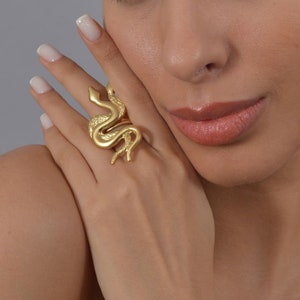 May include: A gold-toned ring featuring a detailed snake design. The ring is worn on a person's finger.
