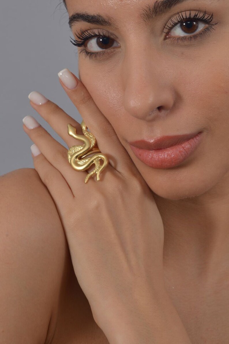 May include: A gold-toned ring featuring a detailed snake design. The ring is worn on a person's finger.