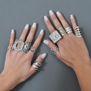 May include: A pair of hands wearing a variety of silver rings. The rings are all different shapes and sizes, and some have intricate designs. The rings are worn on different fingers, and some are stacked on top of each other.
