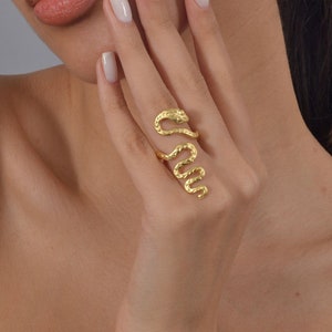 May include: A gold-toned snake ring with a textured finish. The ring is worn on the index finger of a hand with white nail polish.