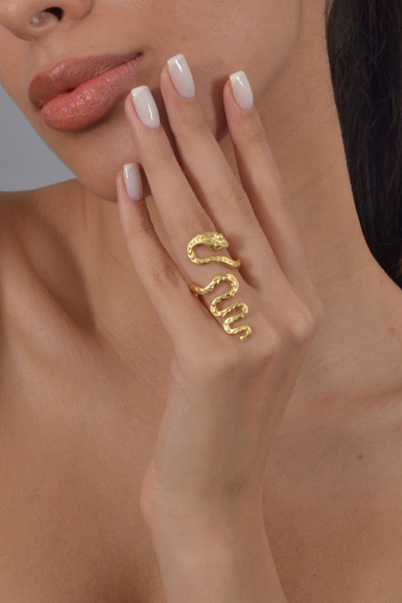 May include: A gold-toned snake ring with a textured finish. The ring is worn on the index finger of a hand with white nail polish.