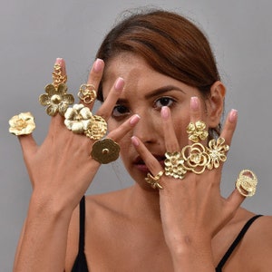 Könnte beinhalten: Die Hände einer Frau, die mehrere goldene Ringe mit floralen Designs trägt. Die Ringe sind auf jedem Finger übereinander gelegt und weisen komplizierte Details auf.