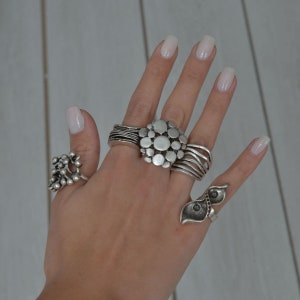 May include: A hand wearing several silver rings. The rings are all different styles, including a ring with a cluster of small circles, a ring with a large circle in the center, and a ring with a leaf design.