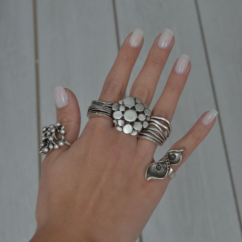May include: A hand wearing several silver rings. The rings are all different styles, including a ring with a cluster of small circles, a ring with a large circle in the center, and a ring with a leaf design.