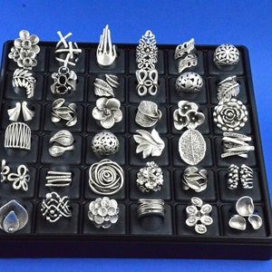 Può includere: Un espositore nero con 36 anelli d'argento con vari disegni, tra cui motivi floreali, geometrici e astratti.