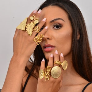 Può includere: Una mano di donna con diversi anelli d'oro. Gli anelli presentano diversi design, tra cui una foglia di felce, una bicicletta, una forma geometrica e un design a spirale.
