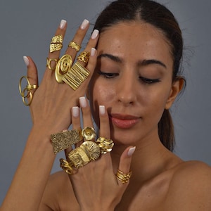 Op de afbeelding: Een vrouwenhand met meerdere gouden ringen met ingewikkelde ontwerpen. De ringen zijn op elke vinger gestapeld en tonen een verscheidenheid aan texturen en vormen.