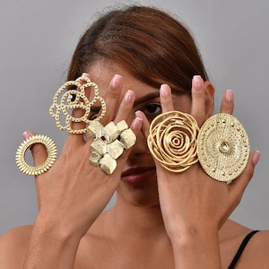 Könnte beinhalten: Die Hände einer Frau, die fünf goldene Ringe trägt. Die Ringe haben alle unterschiedliche Designs, darunter ein Kreis, eine Blume, ein geometrisches Muster und ein Wirbel.