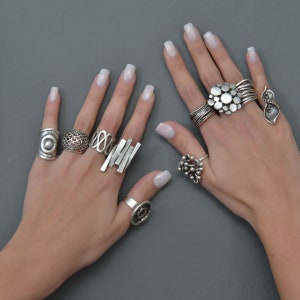May include: A pair of hands wearing multiple silver rings. The rings are of various styles and designs, including a ring with a large, circular design, a ring with a geometric design, and a ring with a floral design.