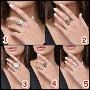 May include: Five different silver rings on a model's hand. The rings are all different styles, including a ring with a cluster of small circles, a ring with a wavy design, and a ring with a simple band.