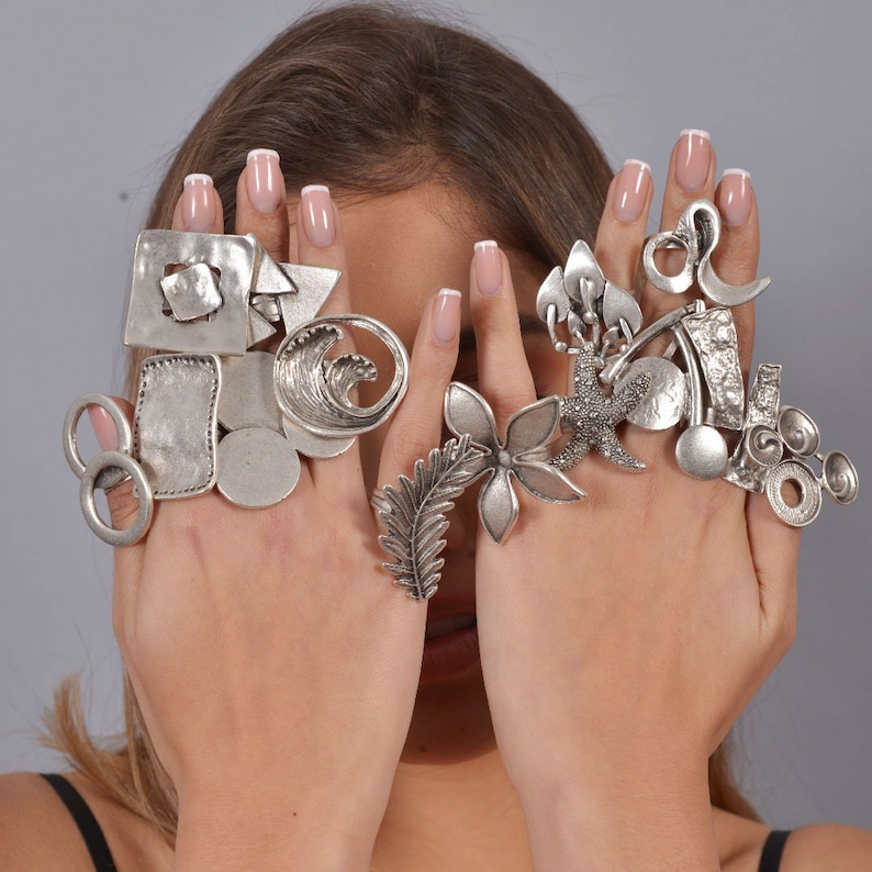 May include: A woman's hands wearing a variety of silver rings. The rings are all different shapes and sizes, and some have intricate designs. The rings are stacked on each finger, creating a bold and eye-catching look.