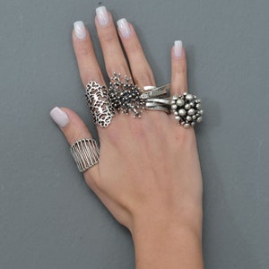 May include: A hand wearing five silver rings. The rings are all different styles, including a delicate filigree ring, a ring with a cluster of small balls, and a ring with a geometric design.