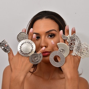 Puede incluir: Un conjunto de anillos de declaración plateados con varios diseños, que incluyen una máscara, una espiral, un círculo con agujeros, un árbol de la vida y un mandala. Los anillos se usan en los dedos de la mano de una persona.
