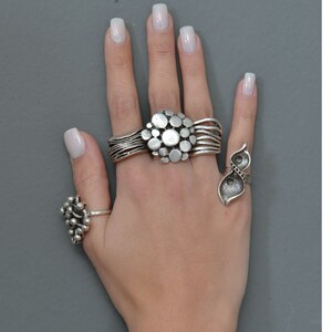 May include: A hand wearing three silver rings. The middle finger has a ring with a cluster of round silver shapes. The ring finger has a ring with two silver leaves. The thumb has a ring with a cluster of small silver balls.