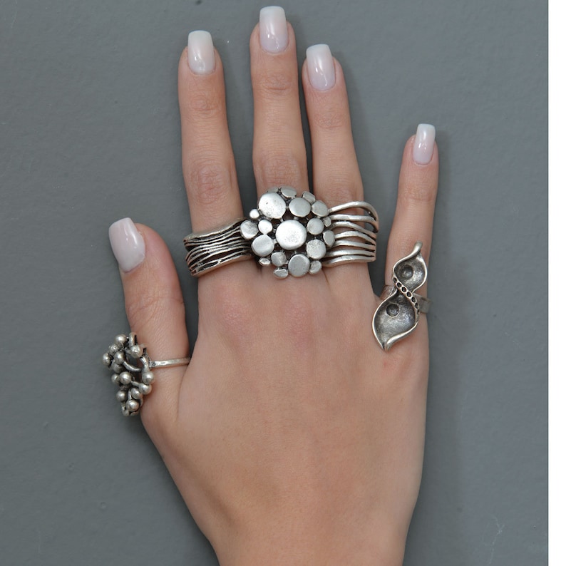 May include: A hand wearing three silver rings. The middle finger has a ring with a cluster of round silver shapes. The ring finger has a ring with two silver leaves. The thumb has a ring with a cluster of small silver balls.