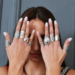 Può includere: Le mani di una persona che indossa diversi anelli d'argento con disegni intricati. Gli anelli sono indossati su dita diverse e presentano varie forme, tra cui motivi floreali, geometrici e astratti.