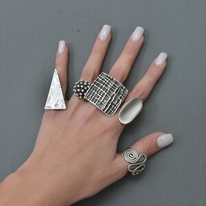 May include: A hand model wearing five silver rings. The rings are all different shapes and sizes. The rings are on the index, middle, ring, and pinky fingers.