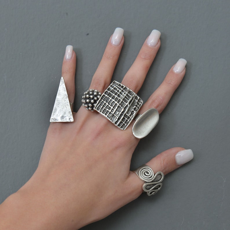 May include: A hand model wearing five silver rings. The rings are all different shapes and sizes. The rings are on the index, middle, ring, and pinky fingers.