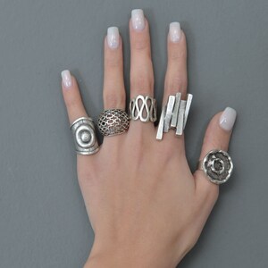 May include: A hand wearing five silver rings. The rings are all different designs, including a circle, a geometric pattern, an infinity symbol, and a stack of rectangles.
