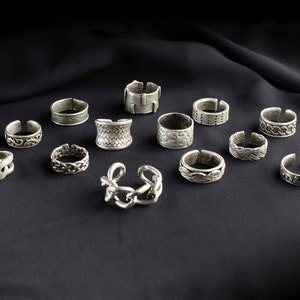 Può includere: Una collezione di 12 anelli d'argento con vari design, tra cui motivi geometrici, maglie a catena e dettagli intricati. Gli anelli sono disposti su uno sfondo di tessuto nero.