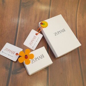 May include: Three white boxes with the word "ZUMABI" printed on them. The boxes are on a wooden surface. One box has a small orange felt flower with a red ladybug on it.