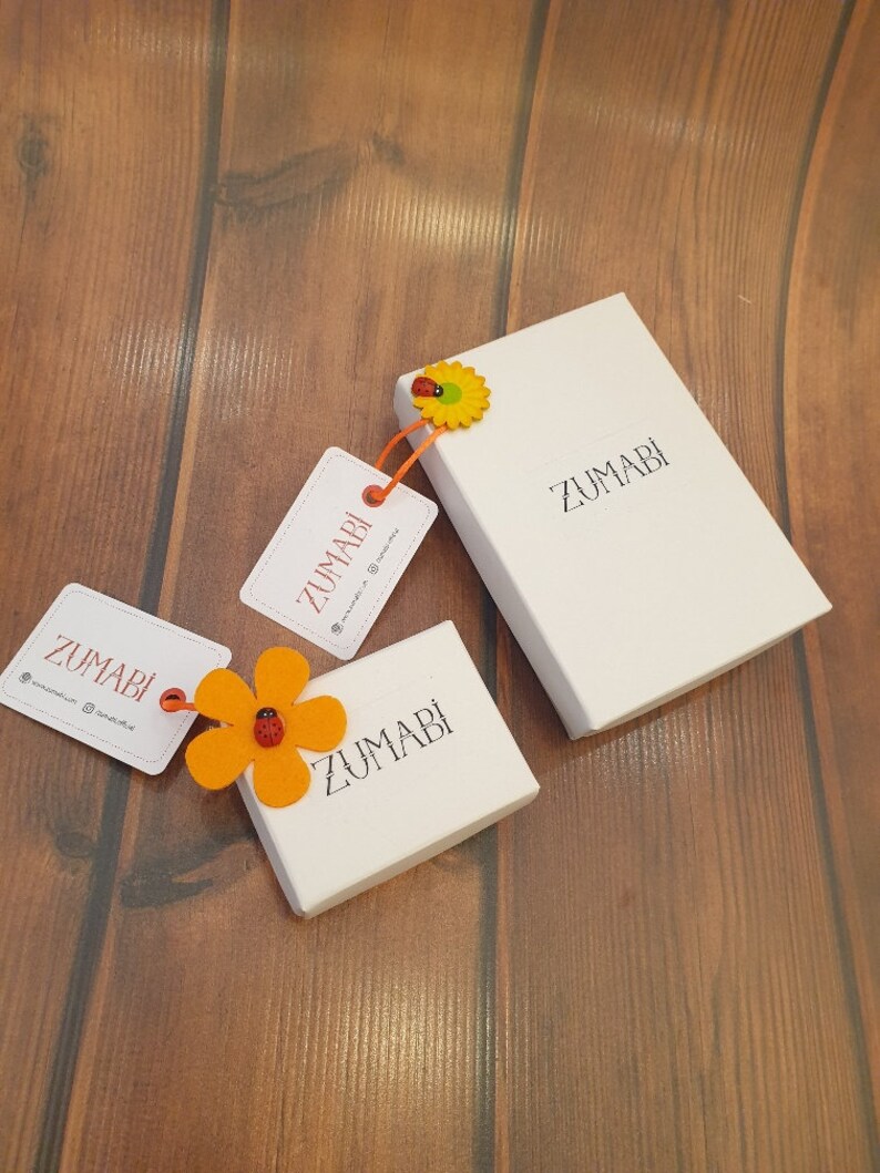 May include: Three white boxes with the word "ZUMABI" printed on them. The boxes are on a wooden surface. One box has a small orange felt flower with a red ladybug on it.