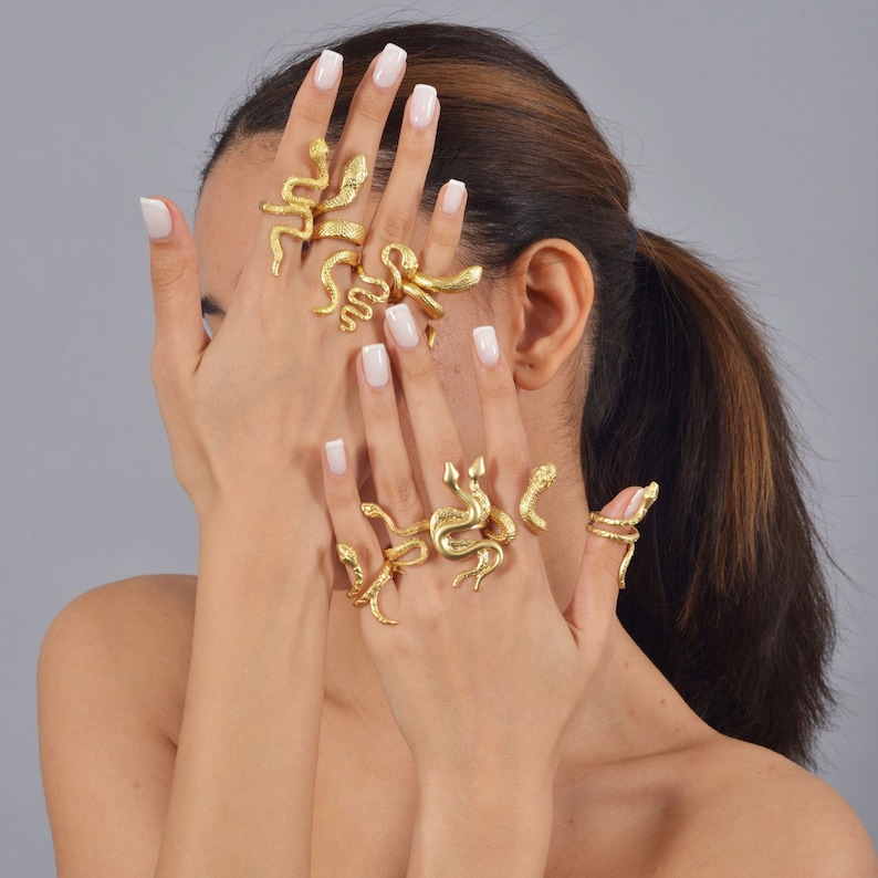 May include: A set of gold snake-shaped rings worn on a person's fingers. The rings are stacked on each other and are a popular fashion accessory.