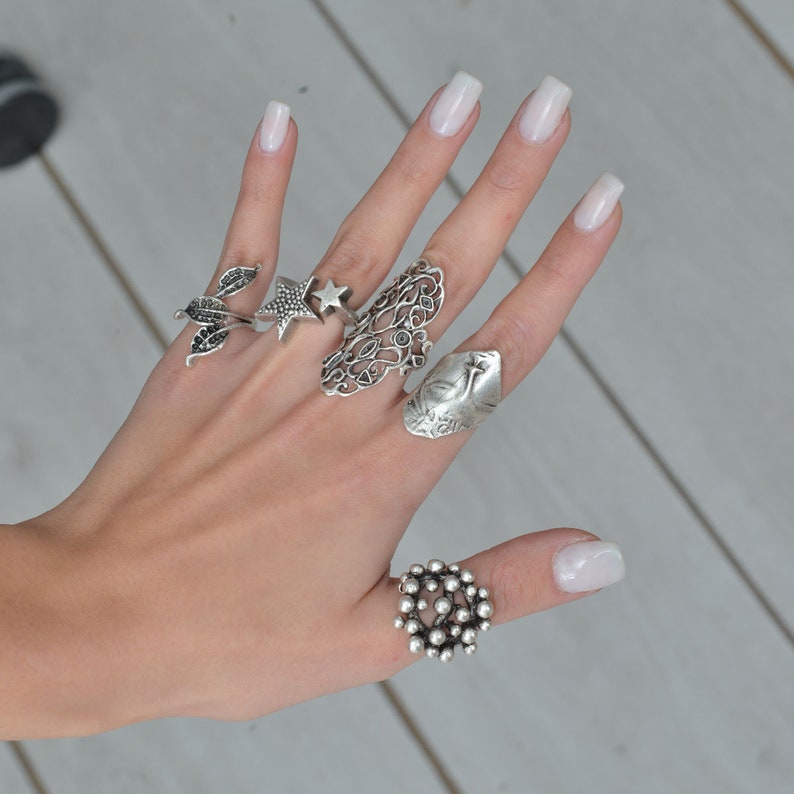 May include: A hand wearing five silver rings. The rings are a star, a leaf design, a floral design, a bird design, and a ring with a cluster of small balls.