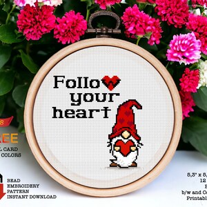 Op de afbeelding: Een ronde borduurring met witte Aida-stof en een rode en witte dwerg die een rood hart vasthoudt. De tekst "Follow your heart" is geborduurd met zwarte draad. De ring is 13,5 cm bij 13,7 cm en is voor 14-count Aida-stof.