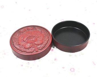 May include: A red, round, lacquered box with a floral design carved into the lid. The lid is separated from the black interior base. The box is likely used for storage or display of small items.