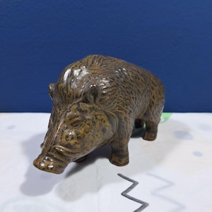 Puede incluir: Figura de cerámica de un jabalí, con un acabado texturizado marrón y beige. El jabalí está representado de pie, con la cabeza baja, mostrando rasgos faciales detallados y un pelaje erizado. Es un objeto decorativo.